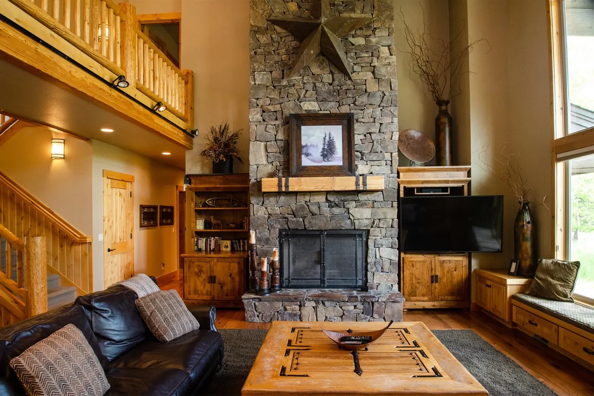 The image showcases the inviting interior of a spacious great room featuring a grand stone fireplace, rich wooden furnishings, large windows framing the outdoors, and a whimsical star decoration. The space is bathed in natural light, creating a warm and welcoming atmosphere ideal for gathering, relaxing, and socializing.