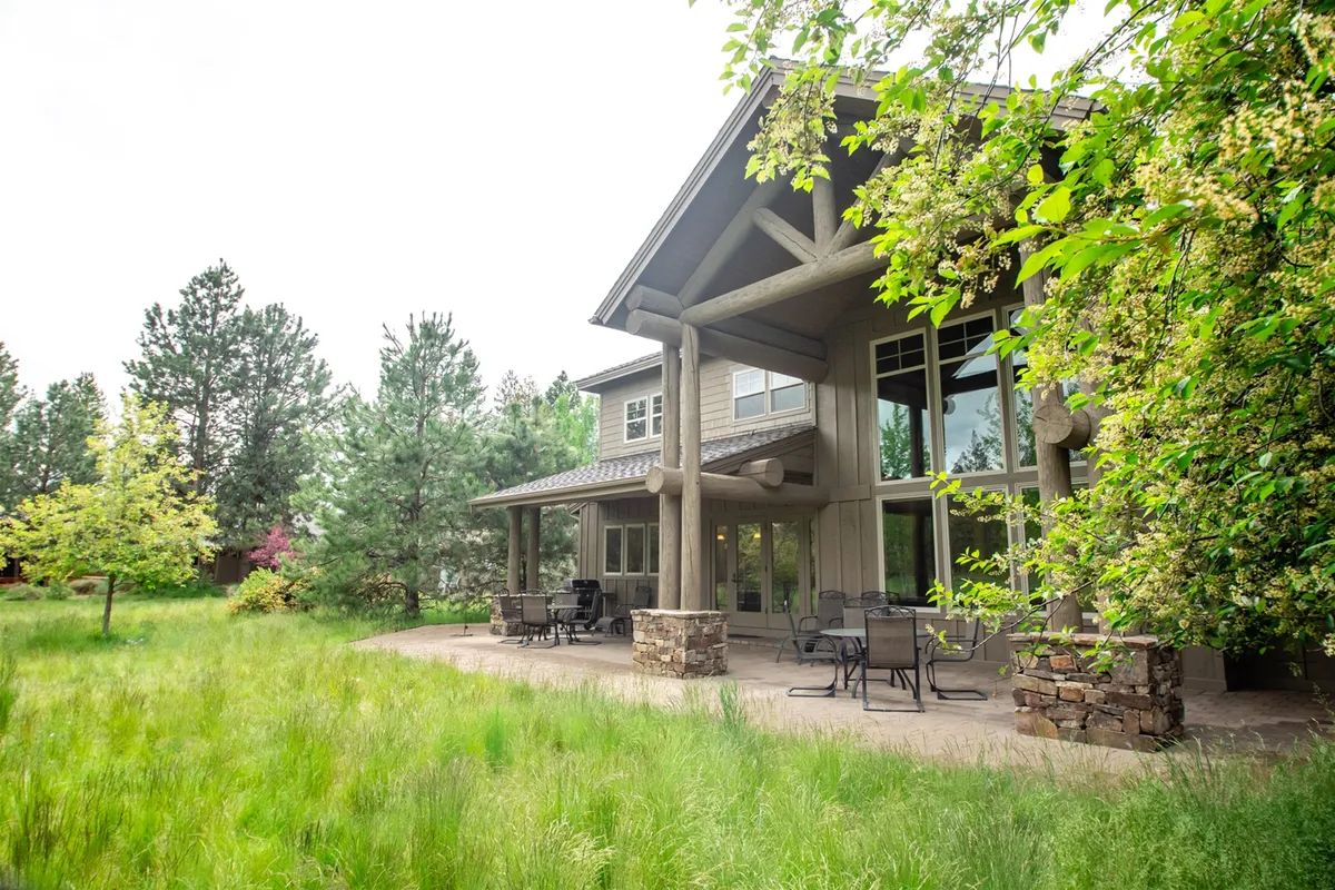 A beautiful exterior view highlighting a covered patio with log pillars, large windows, and an outdoor seating area. Surrounded by natural landscaping, the patio is set up for relaxation and socializing, with clear sightlines to the surrounding scenery, inviting guests to unwind and soak up Sunriver's peaceful ambiance.