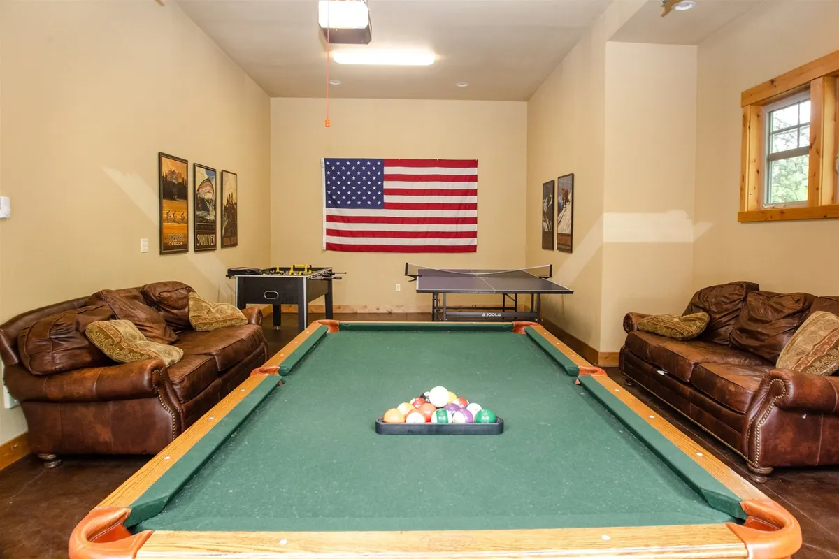 This image captures a game room with a pool table, ping pong table, comfortable sofas, and an American flag on the wall. The scene radiates a relaxed and entertaining mood, perfect for gatherings and games with friends or family. Ample seating and the multiple game tables are clearly visible, emphasizing the space's versatility and fun atmosphere.