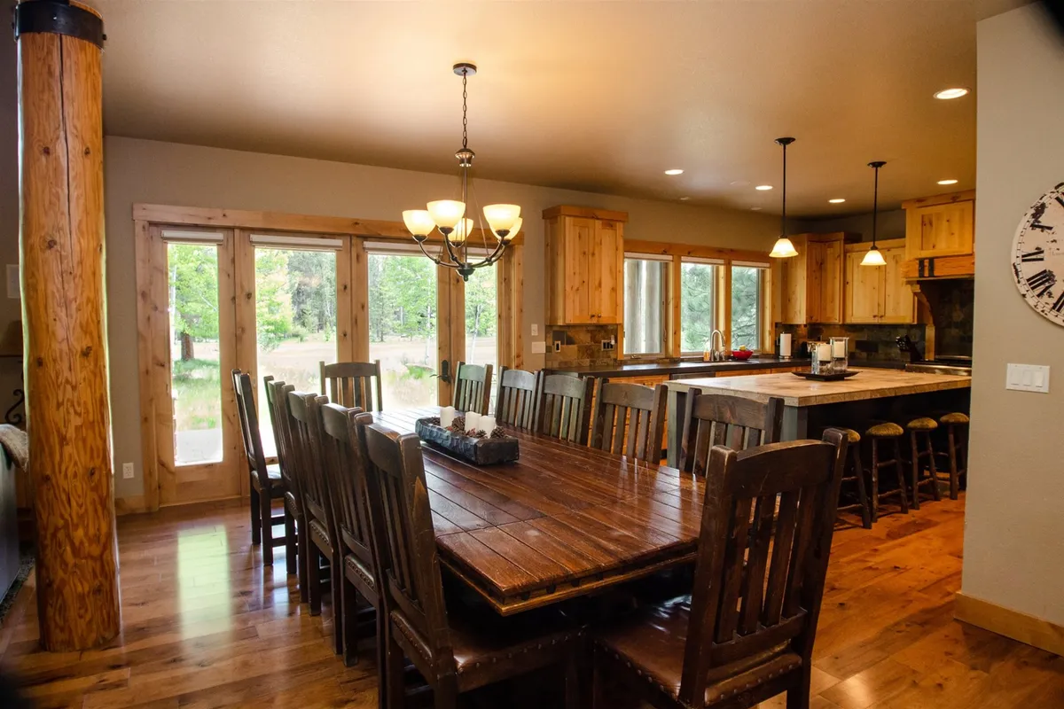 A large wooden dining table sits beneath a stylish chandelier, flanked by a kitchen island and surrounded by cabinetry and large windows. The setting radiates warmth and community, inviting guests to gather for meals, celebrations, and memorable shared moments.