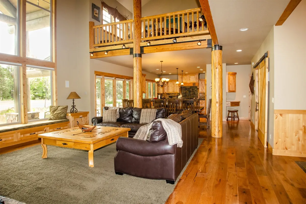 This image features a spacious open-plan great room with large windows that let in abundant natural light. Wooden beams accent the ceiling, while plush, comfortable seating is arranged near a roaring fireplace. The dining area is seamlessly connected, creating an inviting space for family gatherings, reading by the window, or enjoying cozy evenings by the fire. The view through the windows hints at a beautiful Oregon landscape beyond.